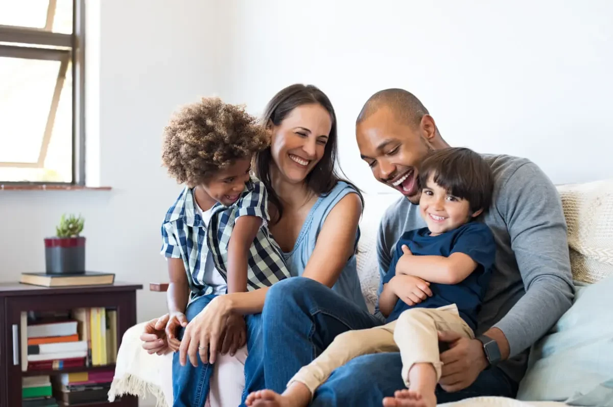 Happy Family sitting on a bench in a modern interior, wearing a beige sleeveless top and brown high-waisted pants, with sunlight casting a shadow on the wall behind her.