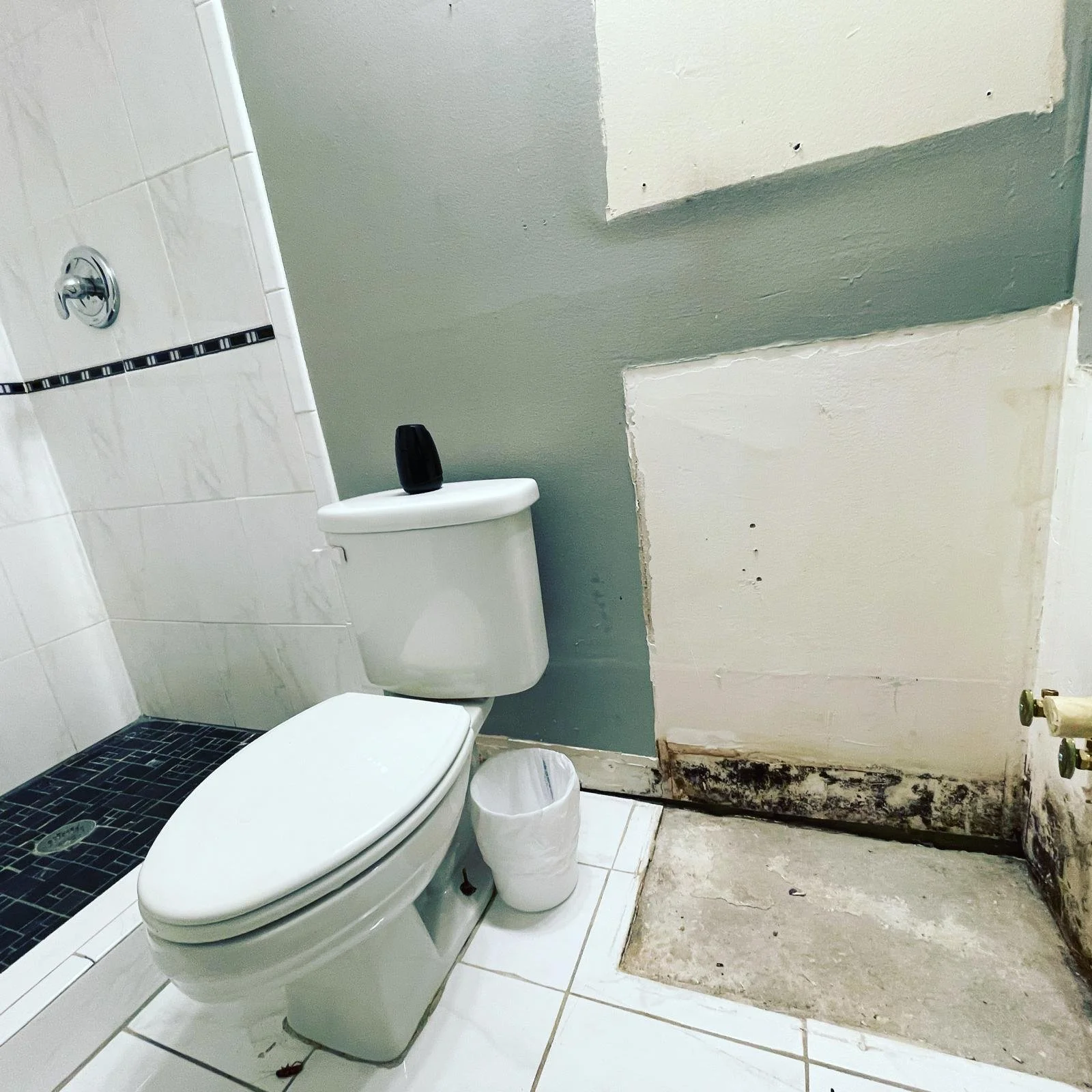 A bathroom with a toilet, a trash can, a partially finished wall, and a shower area with black tile and a silver shower handle.