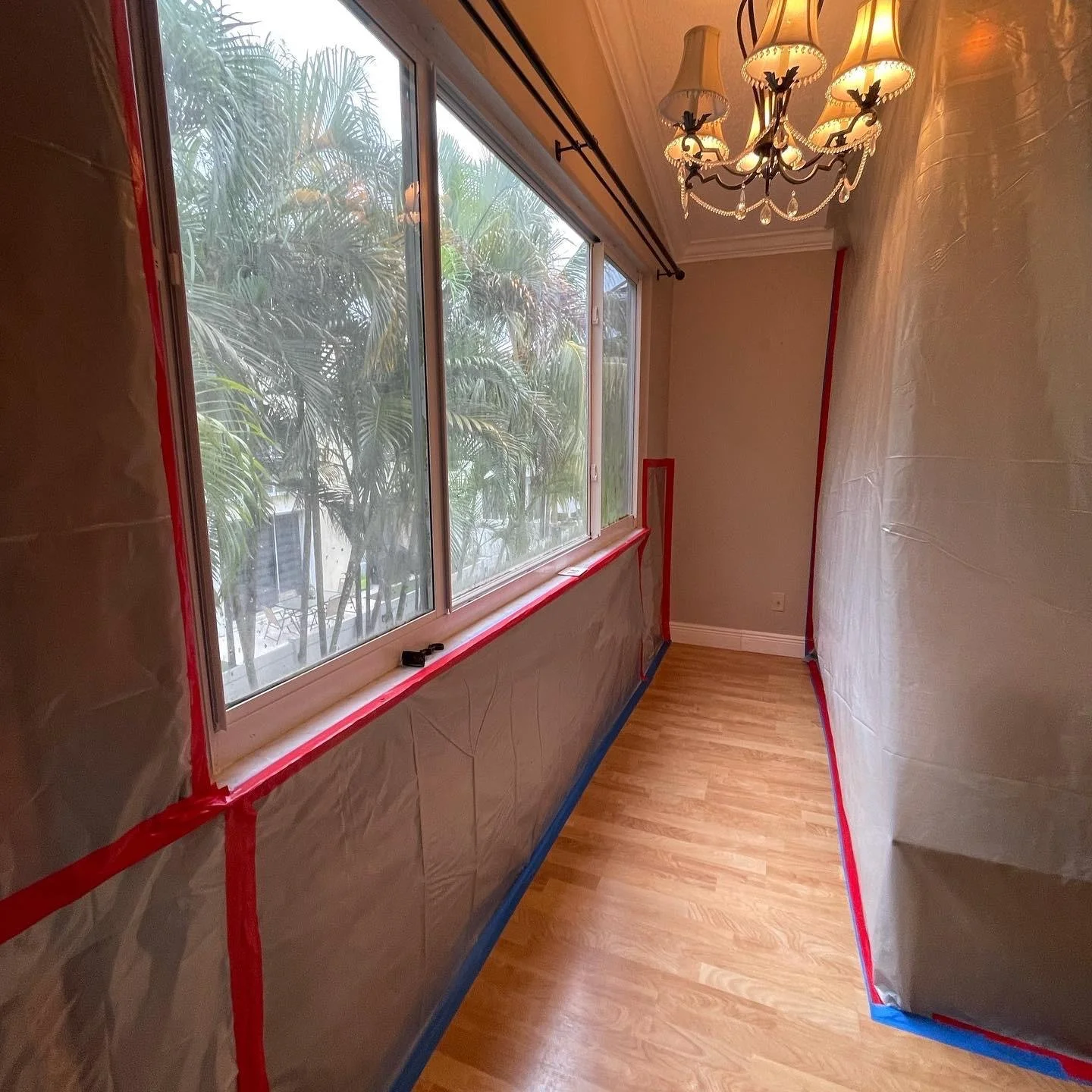 A narrow room with plastic sheeting and painter's tape on the walls, wooden flooring, a large window with a view of green trees, and an ornate chandelier hanging from the ceiling.