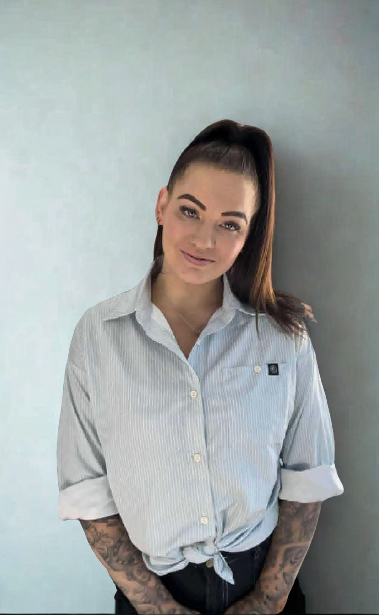 A woman with a high ponytail hairstyle, wearing a light striped button-up shirt and black pants, standing against a plain gray wall.