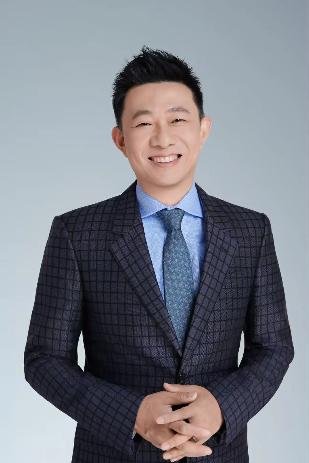 A smiling man dressed in a dark plaid suit with a light blue shirt and patterned tie, standing with hands clasped against a plain, light gray background.