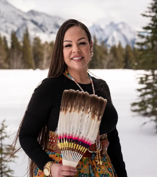 Woman smiling outdoors in winter, holding a fanned collection of feathers, with snowy landscape and mountains in the background.