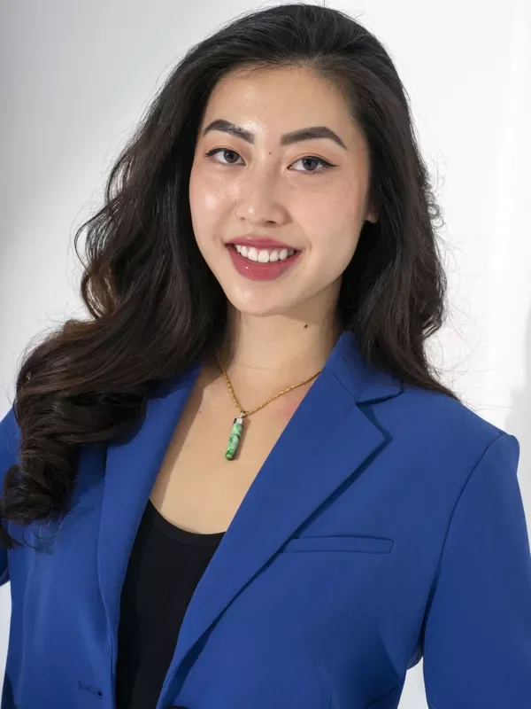 A young woman with long, wavy dark hair, wearing a blue blazer over a black top, smiling, and wearing a green pendant necklace, standing against a plain white background.