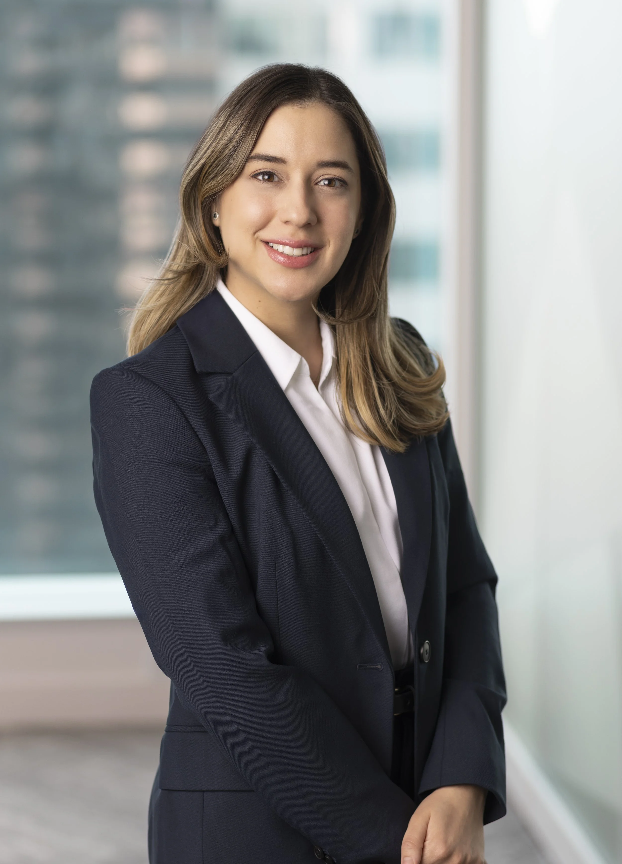Portrait of a young woman in a business suit smiling, standing indoors near a large window with a blurred cityscape in the background.