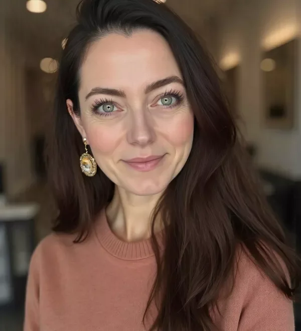 A woman with long dark hair, light skin, and blue eyes, wearing a pinkish-brown sweater and ornate earrings, smiling softly inside a warmly lit room.