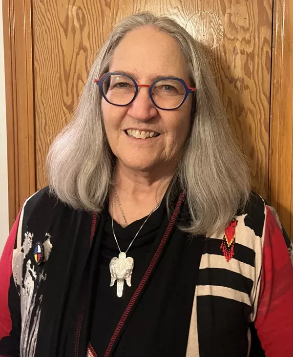 A smiling older woman with long gray hair, blue and red glasses, wearing a black top, layered necklaces, and a colorful jacket with black, white, red, and pink patterns, standing in front of a wooden wall.