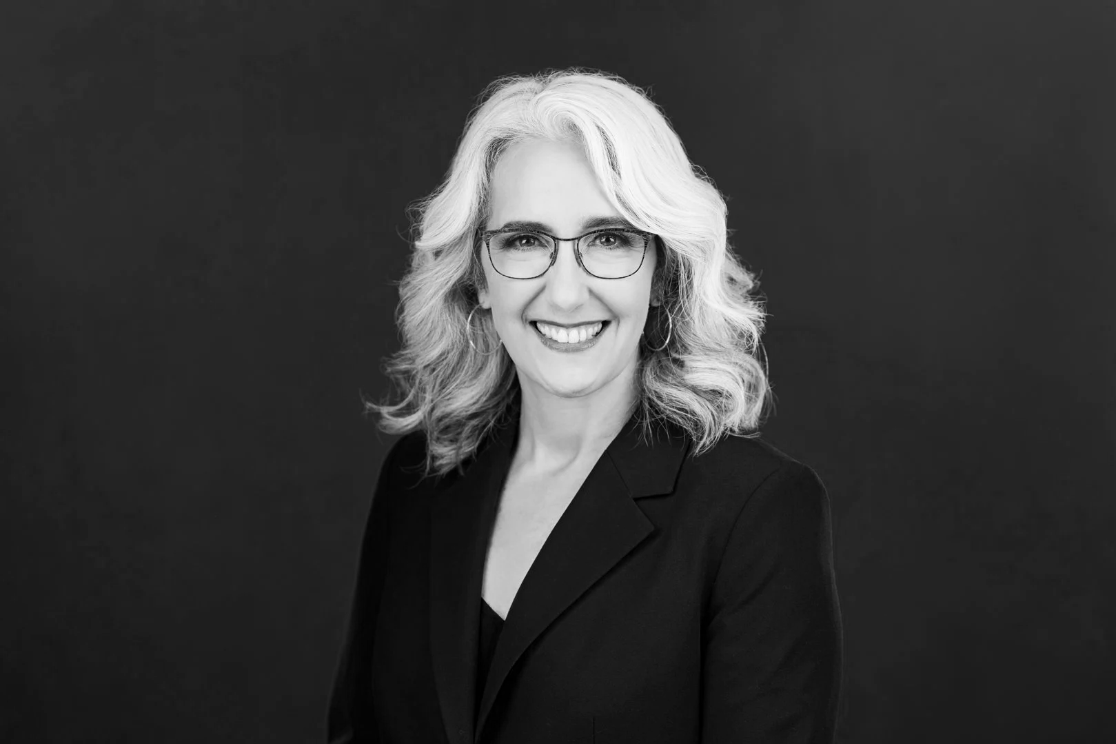 Cori Ghitter smiling with glasses and wavy shoulder-length hair, wearing a dark blazer, against a dark background.