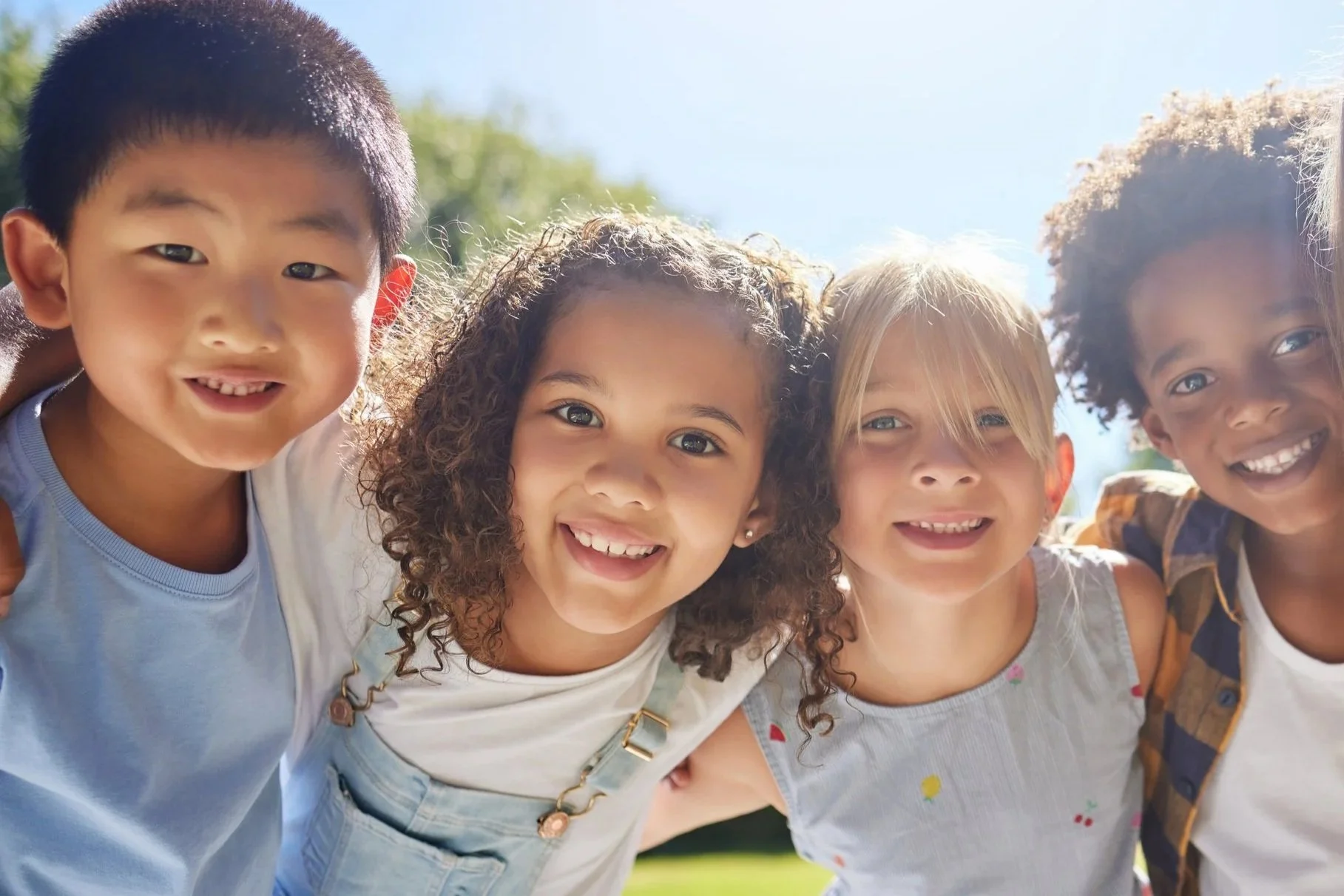 Diverse group of five smiling children standing close together outdoors on a sunny day.