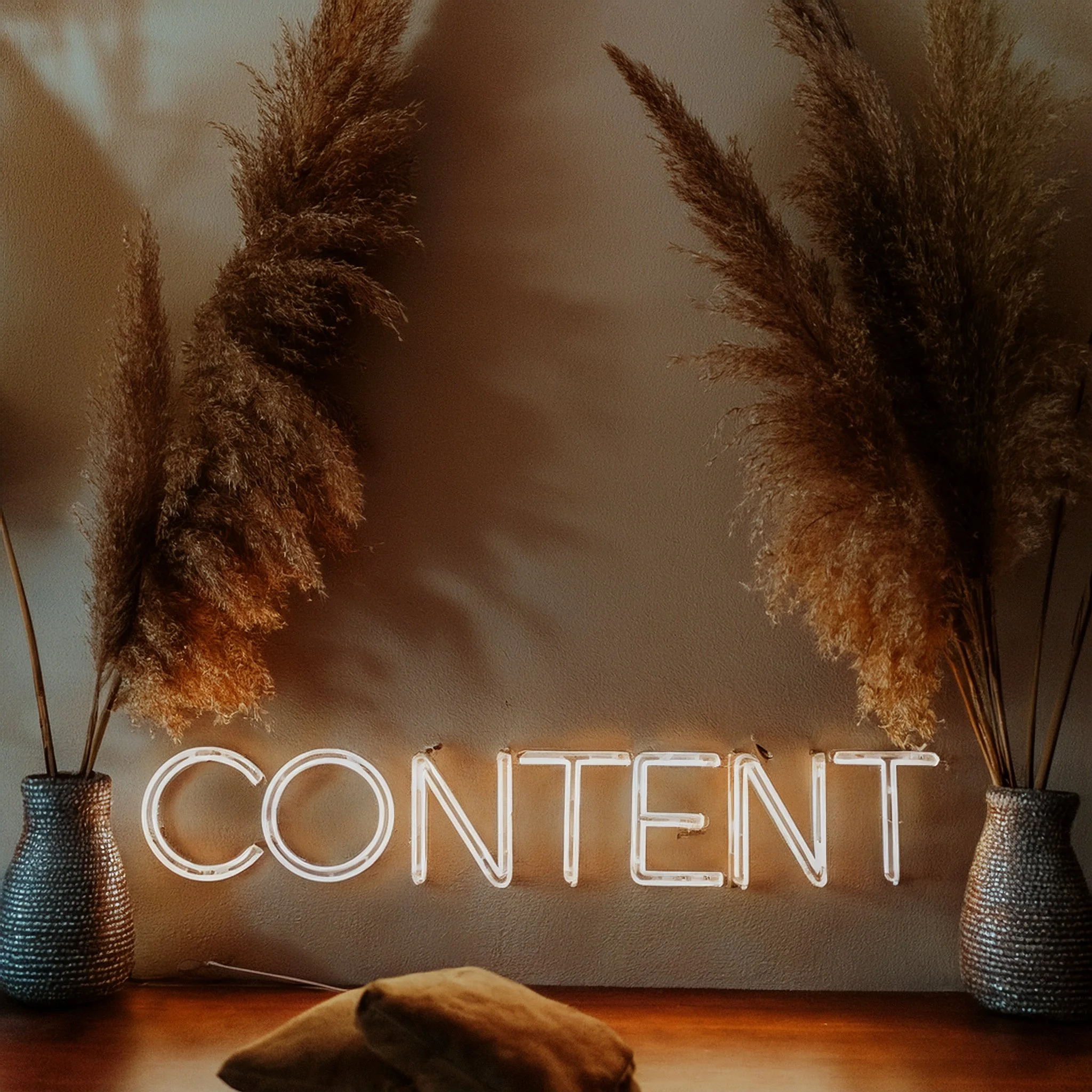 Decorative wall with illuminated neon sign that spells out 'CREATIVE', flanked by two tall vases filled with dried pampas grass, on a wooden table.