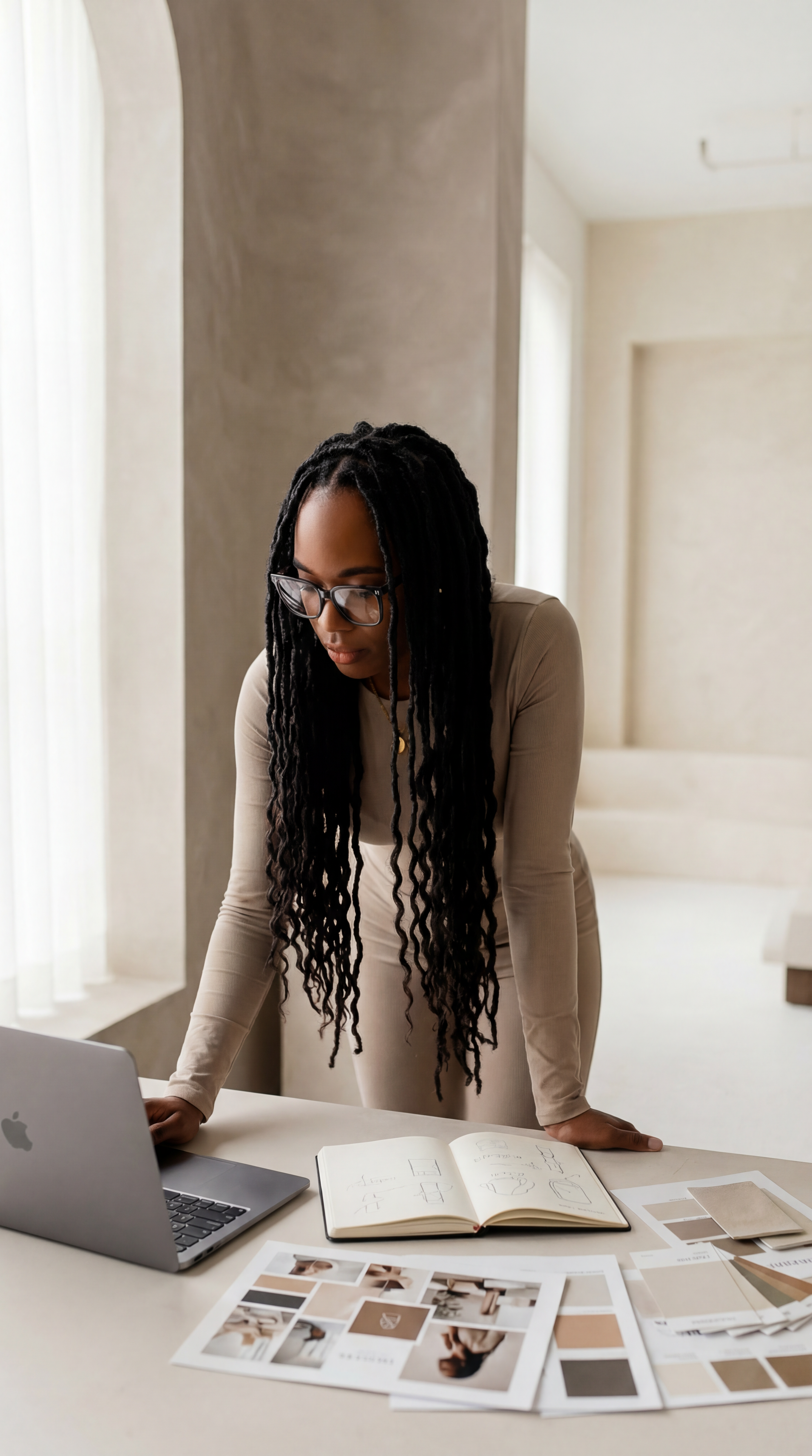 A woman with long dreadlocks and glasses leaning over a table with design sketches, open book, color samples, and a laptop in a well-lit room.