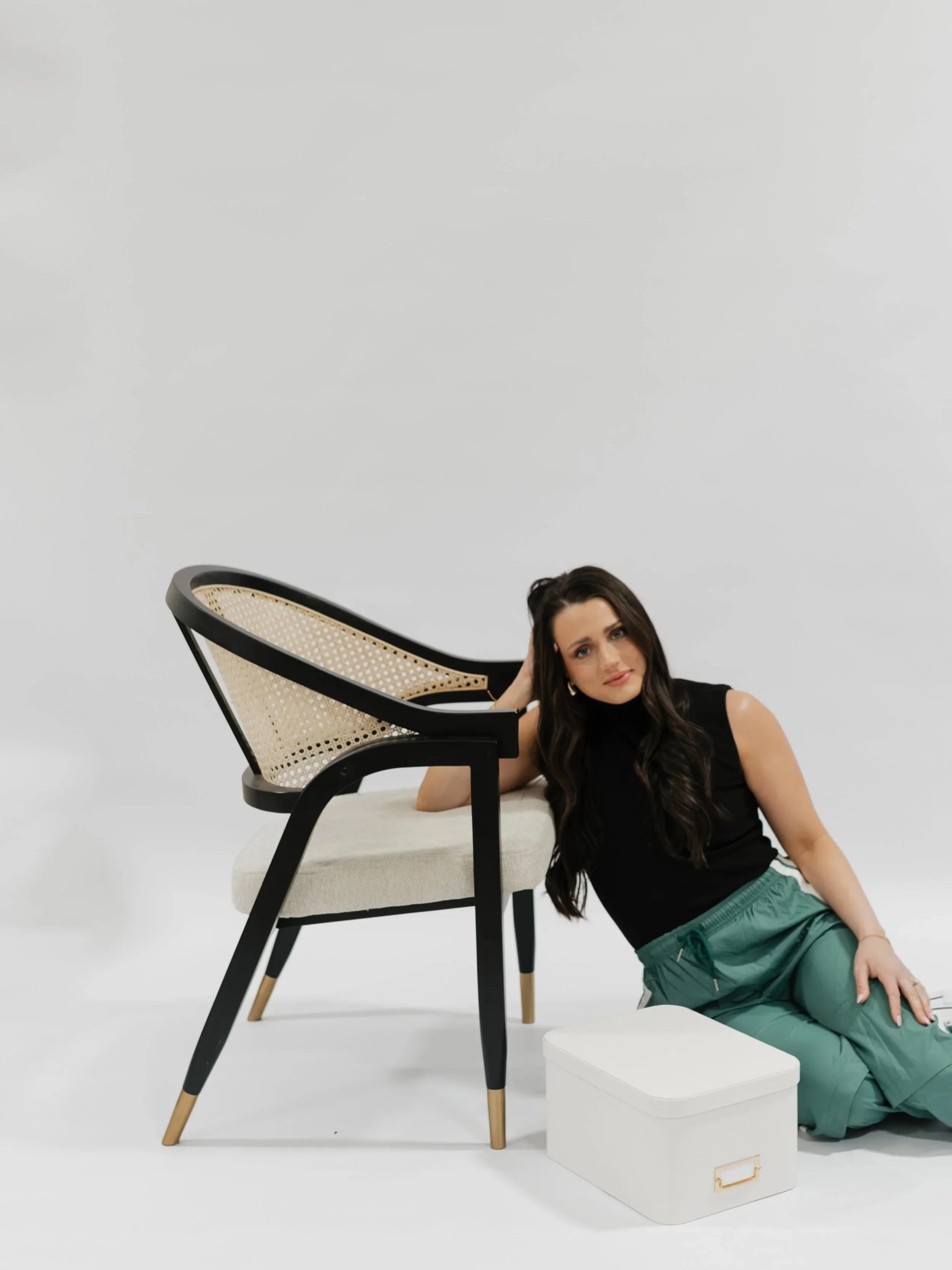 A woman with long dark hair wearing a black sleeveless top and green pants sitting on the floor, leaning against a white bench with a black and beige chair with a cane backrest and wooden legs placed upside down on the white surface near her.