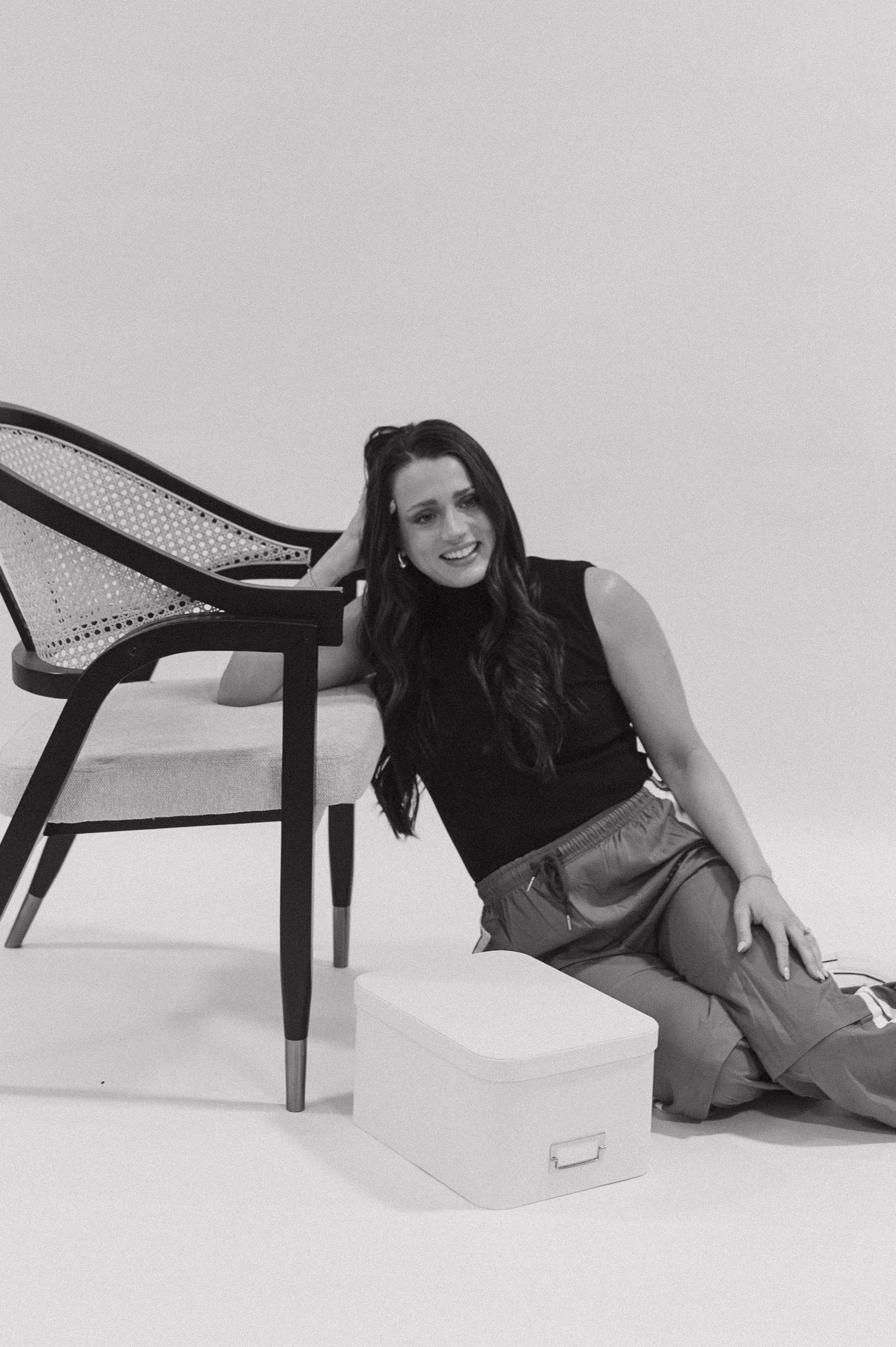 A young woman with long dark hair, wearing a black sleeveless top and casual pants, is sitting on the floor and leaning against a white box in front of a chair. She is smiling and looking slightly to the side, with her right hand resting on her head.