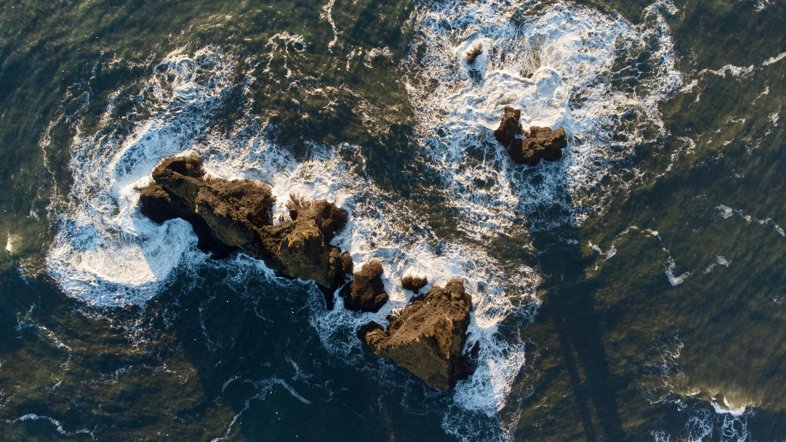Aerial view of ocean waves crashing against rocky formations in the water.