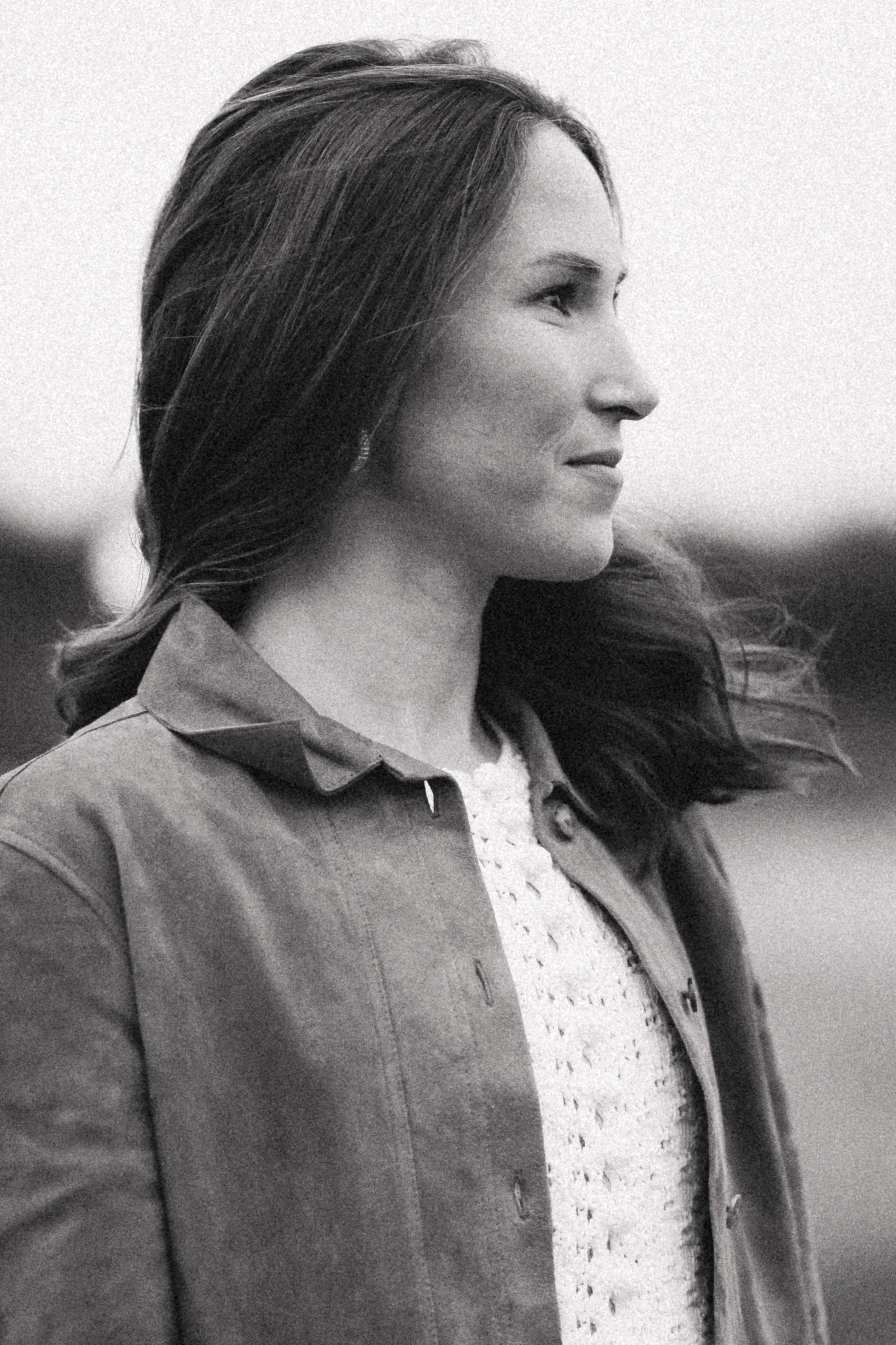 A woman with long dark hair, wearing a collared shirt and a jacket, is looking to the right outdoors in black and white.