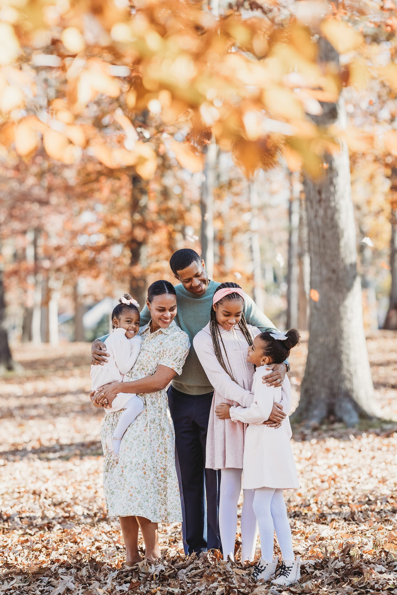 A family of five, consisting of two women, one man, and two young girls, is enjoying a walk in a park during autumn, surrounded by trees with orange leaves.