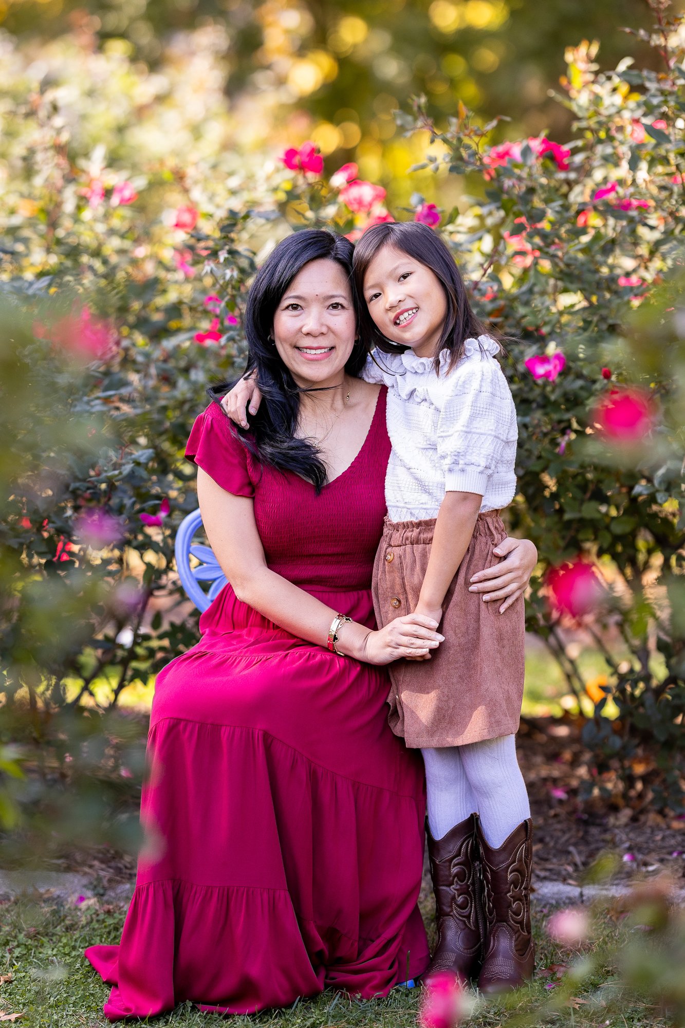 A woman and a young girl pose together outdoors surrounded by pink and red flowers. The woman is wearing a red dress, and the girl is in a white blouse with brown skirt and cowboy boots. They are smiling and embracing in front of blooming bushes.