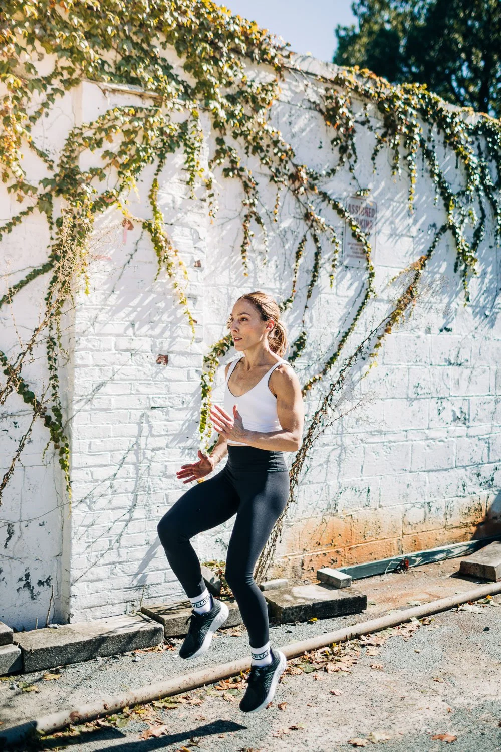 A woman jogging outdoors on a sunny day, wearing a white tank top and black leggings, with a white brick wall and climbing greenery in the background.
