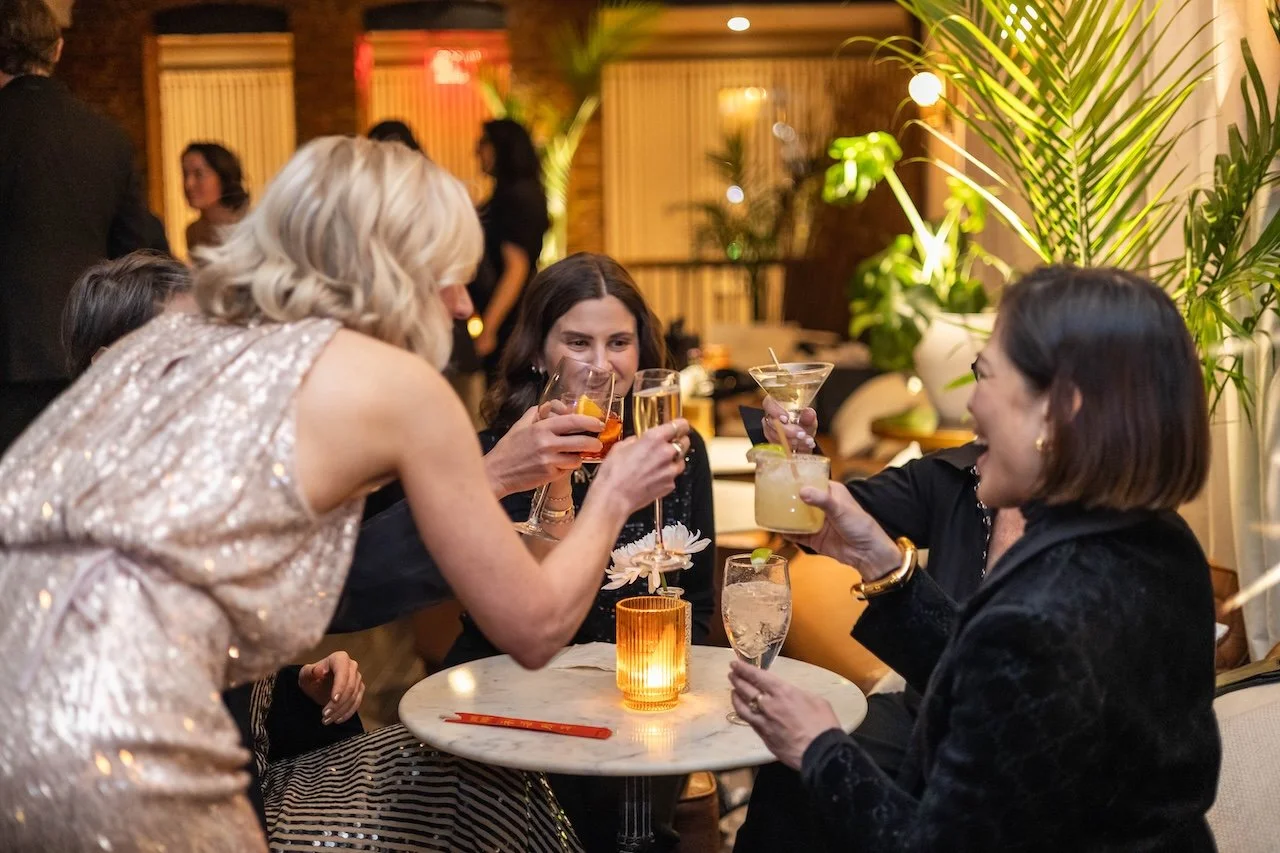 Four women at a bar or restaurant table toasting with drinks, surrounded by indoor plants, with other people in the background.