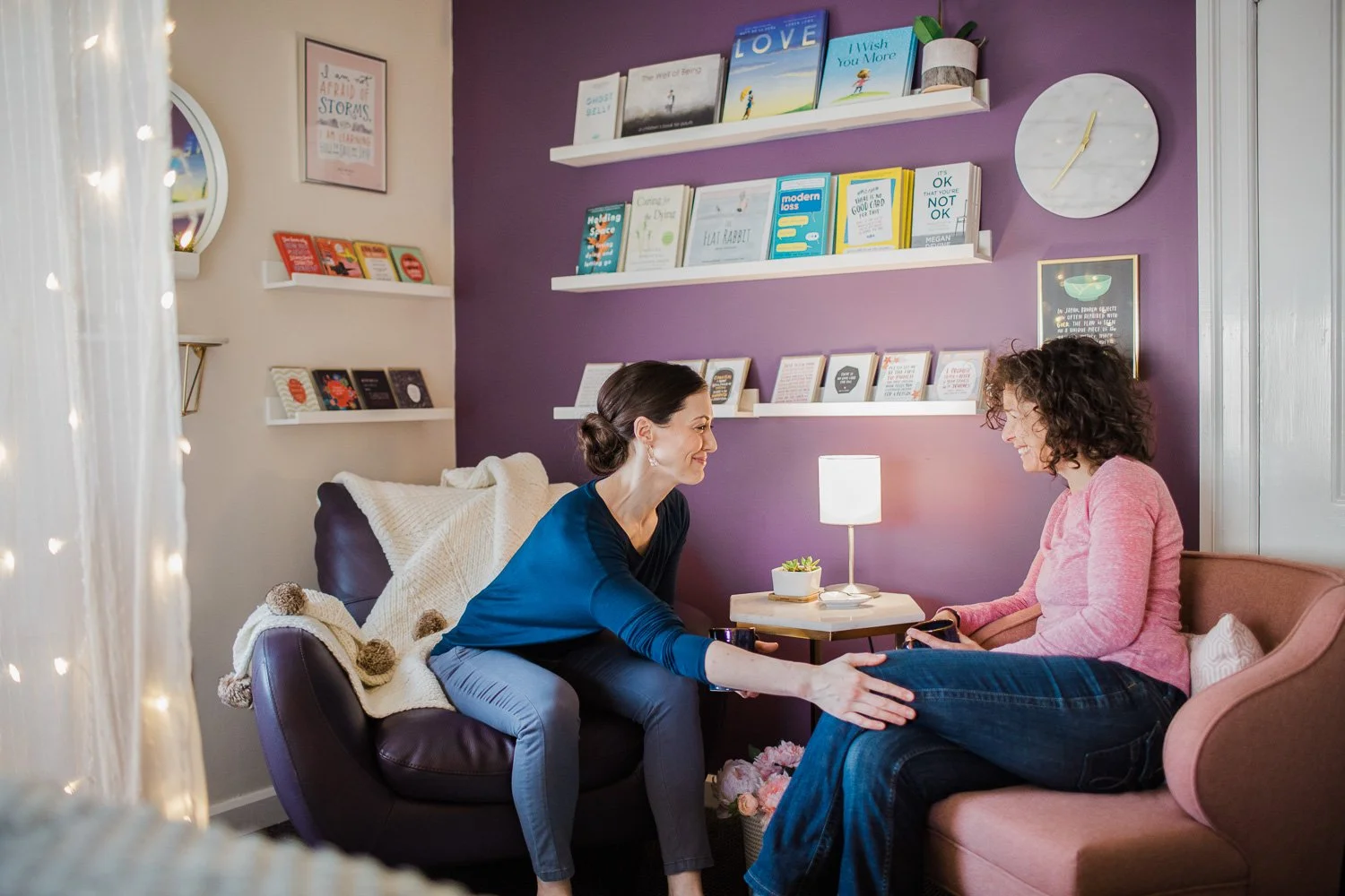 Two women sitting in a cozy living room, smiling and engaging in conversation. One woman is sitting on a purple couch, the other on a pink armchair. The room has a purple and cream wall decorated with books and framed art, a small side table with a l
