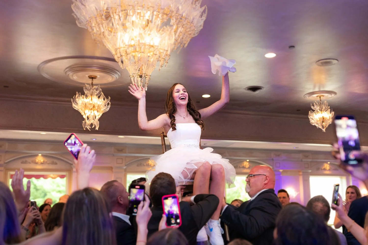 A woman in a white dress is being lifted on a chair during aBat Mitzvah, surrounded by people taking photos, in an elegant decorated room with chandeliers and purple lighting.