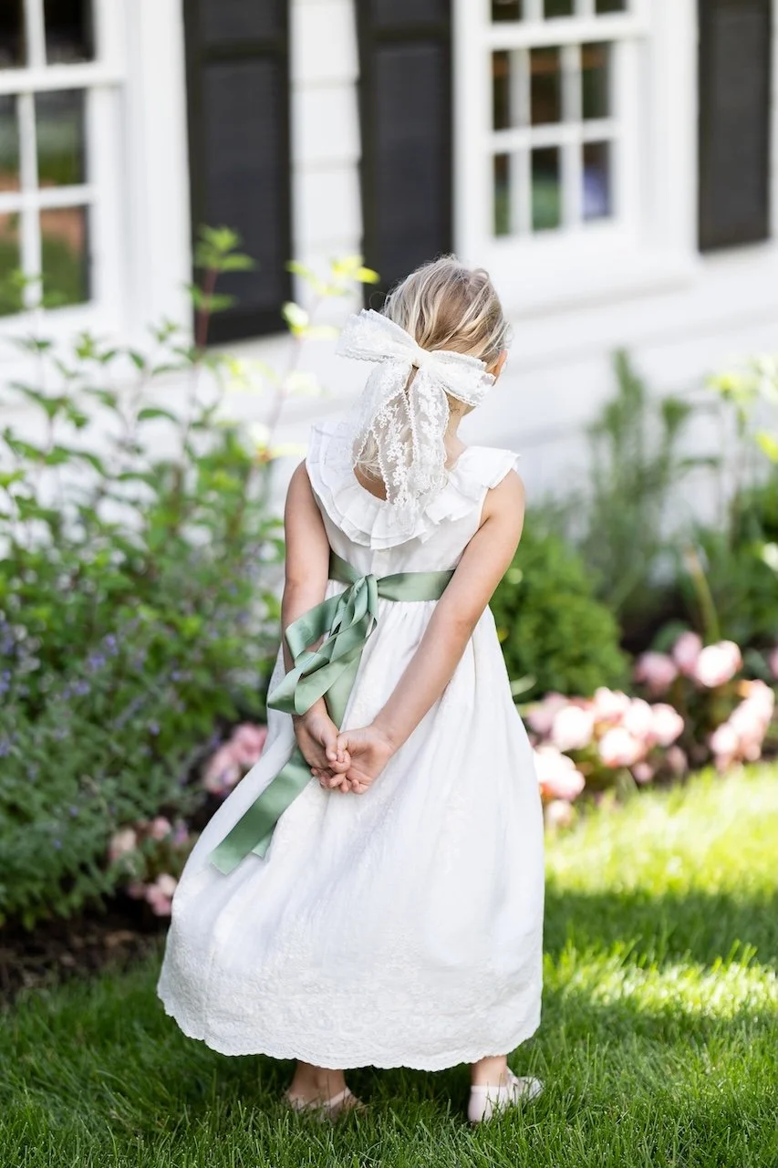 A young girl in a white communion dress with a green sash bow, standing on a lawn in Montclair, N in front of flowering bushes, with her back to the camera and her hands clasped behind her back.