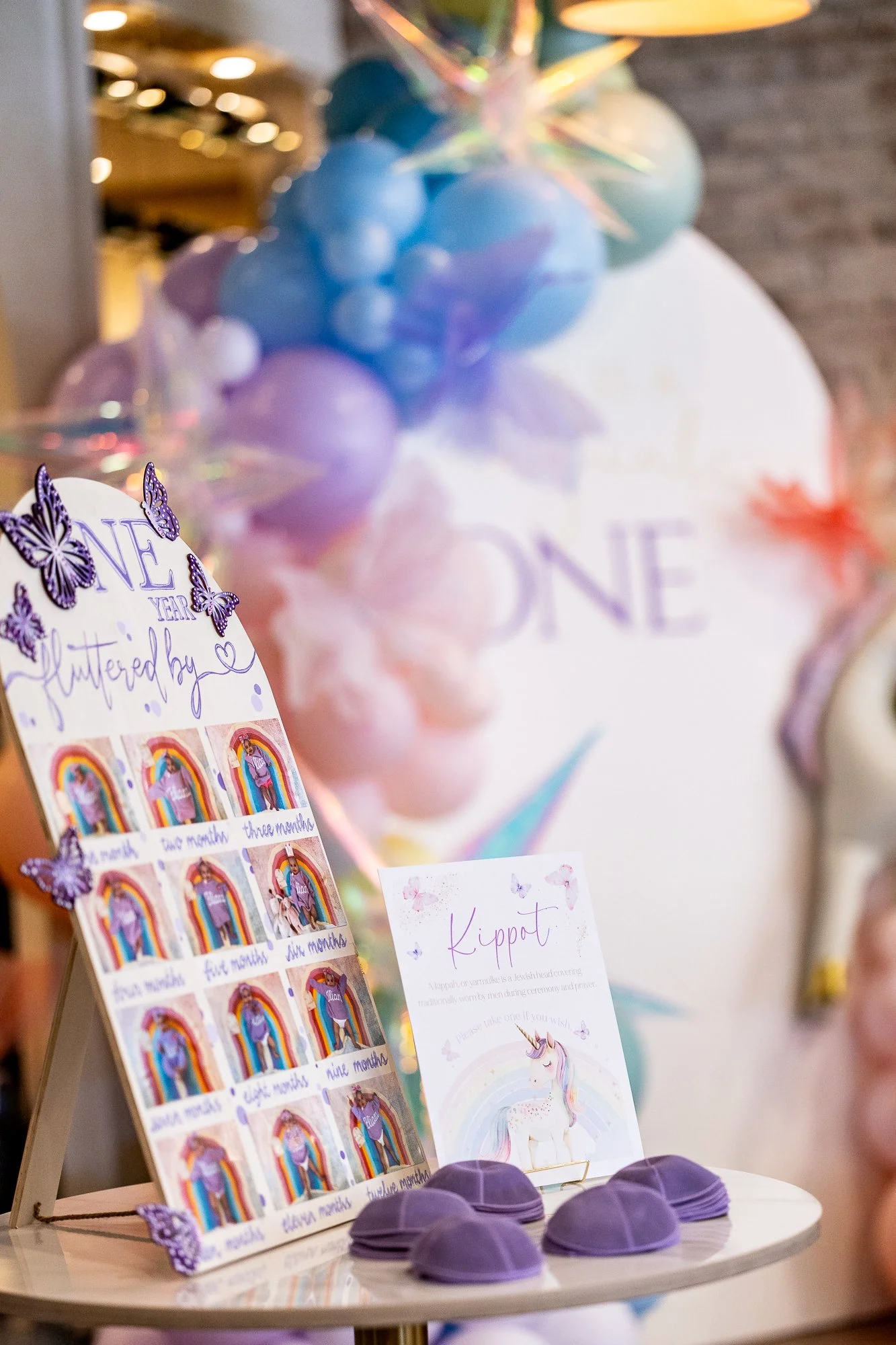 A display with purple hats, a poster about Hanukkah, and a decorative sign with butterflies and a handwritten message. In the background, pastel-colored balloons, including blue, purple, and pink, are arranged in a cluster with a shiny star-shaped de