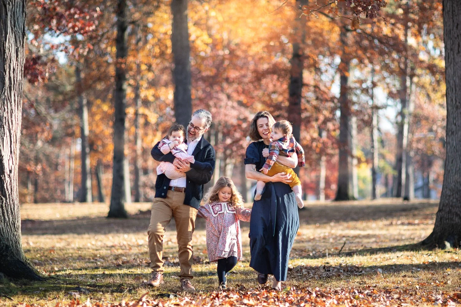 A family of four walking in a park with fall foliage. The father wearing a black jacket carries a baby girl, and the mother in a blue dress holds a toddler. A young girl walks between them, holding hands.
