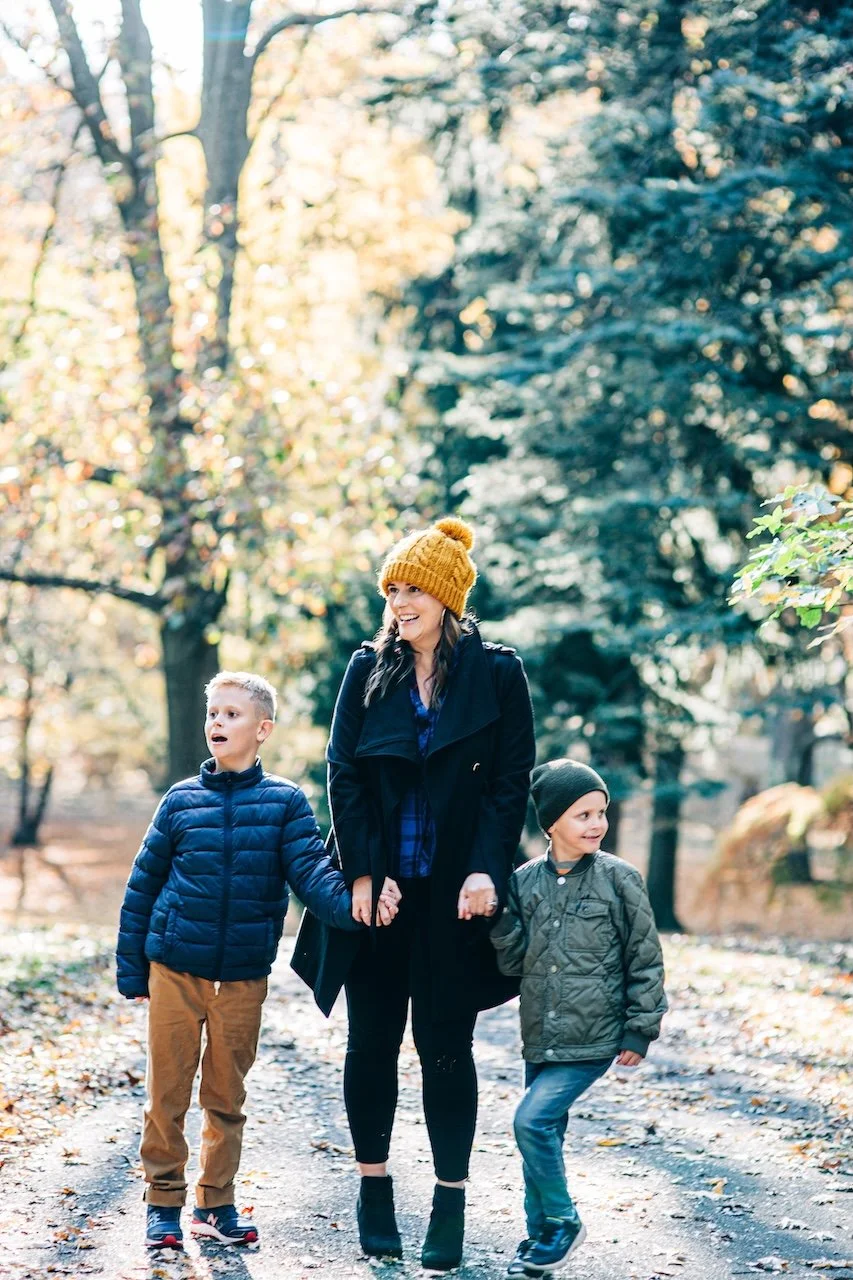 A woman walking with two children in a park during autumn, holding hands with each child. The woman is smiling and wearing a yellow knit hat, black coat, and black shoes. The children are dressed in jackets and hats, with one child in a blue jacket a