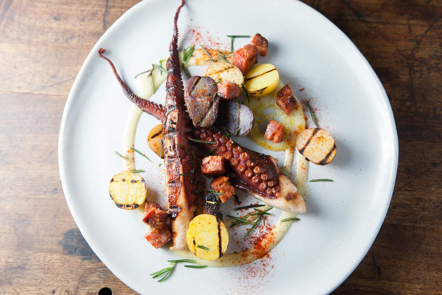 Grilled octopus tentacle served with roasted potatoes, grilled vegetables, herbs, and garnished with spices on a white plate.