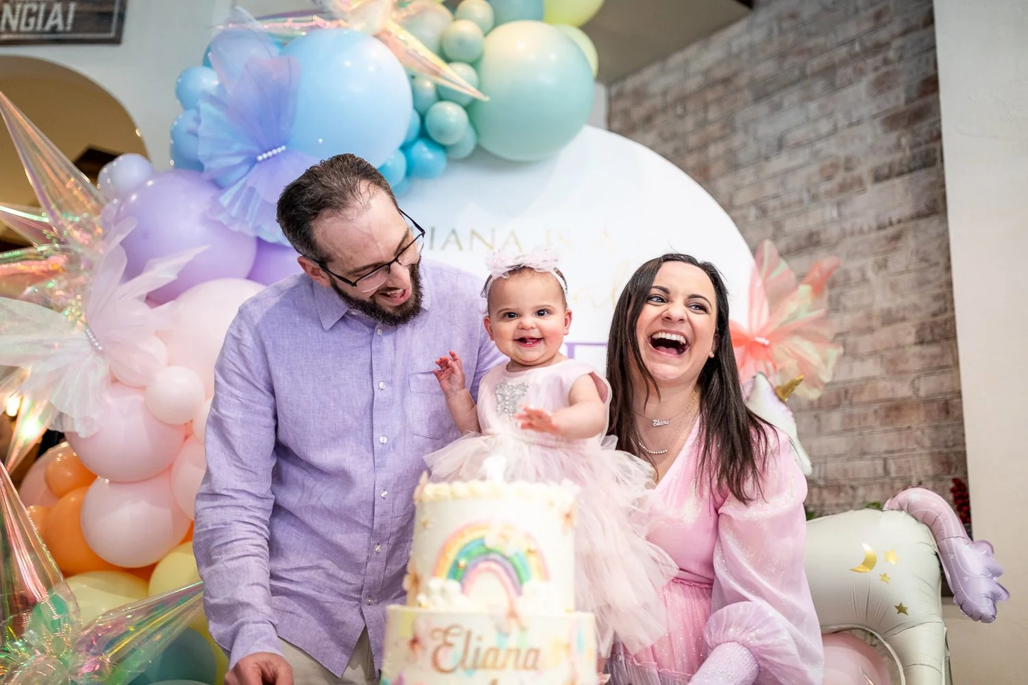 A family celebrating a young girl's 1st birthday with balloons, a decorated cake with a rainbow, and a pink princess dress. The girl is smiling and sitting with her family.