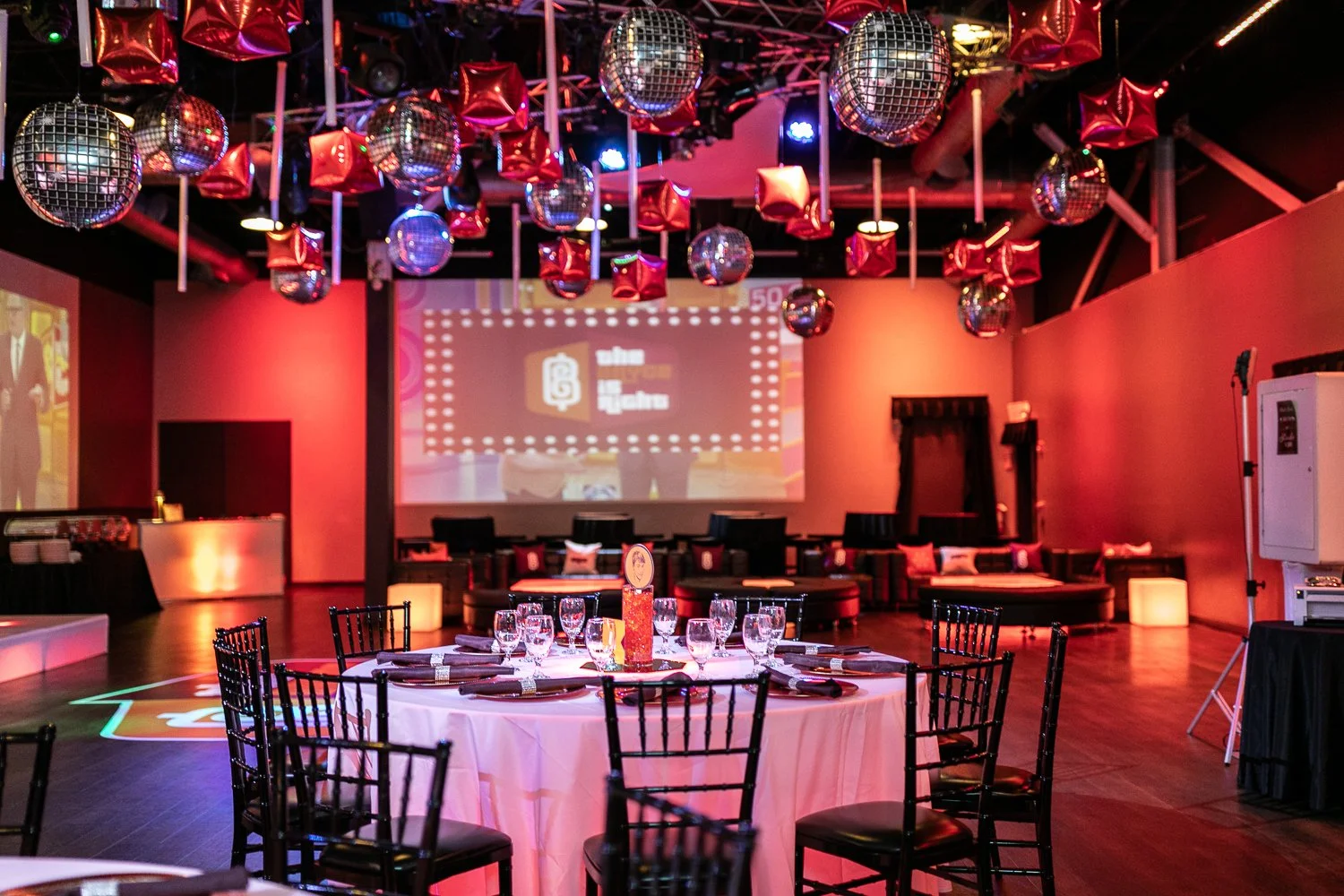 An event space with a large screen on the back wall displaying a logo, surrounded by red and silver balloons and disco balls hanging from the ceiling. The room has tables set with glassware and black chairs, with soft lighting creating a festive atmo