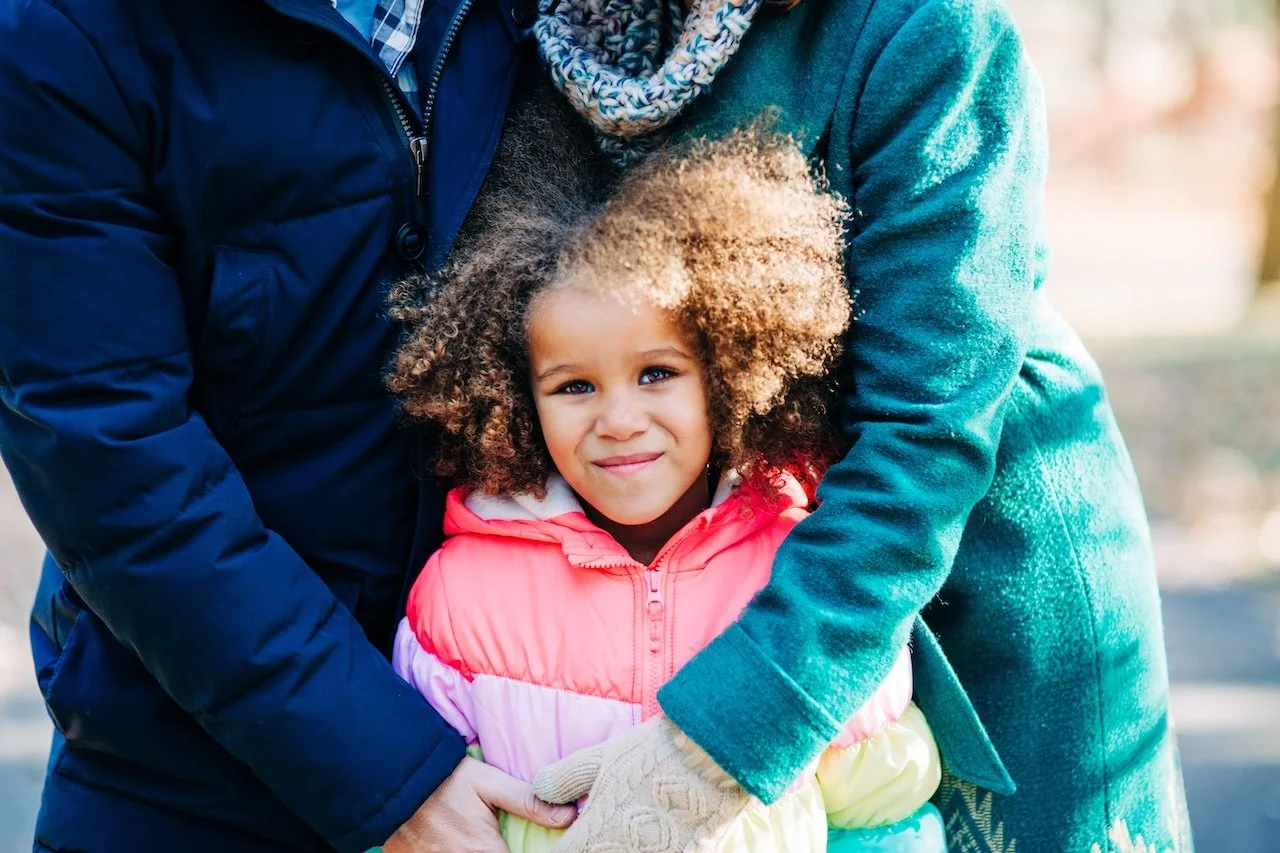 A young girl with curly hair wearing a pink and purple jacket being hugged by two adults. One adult wears a blue jacket and the other a green jacket. The scene takes place in Brookdale Park, NJ for a fall mini session.