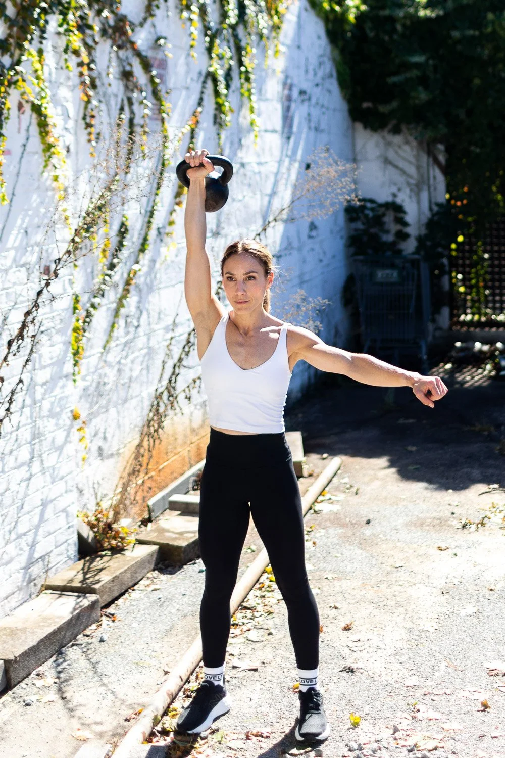 Woman performing an overhead kettlebell exercise outdoors, wearing a white tank top and black leggings.