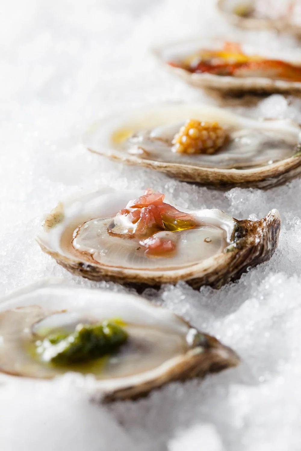 Close-up of fresh oysters on a bed of ice, topped with various sauces and garnishes.