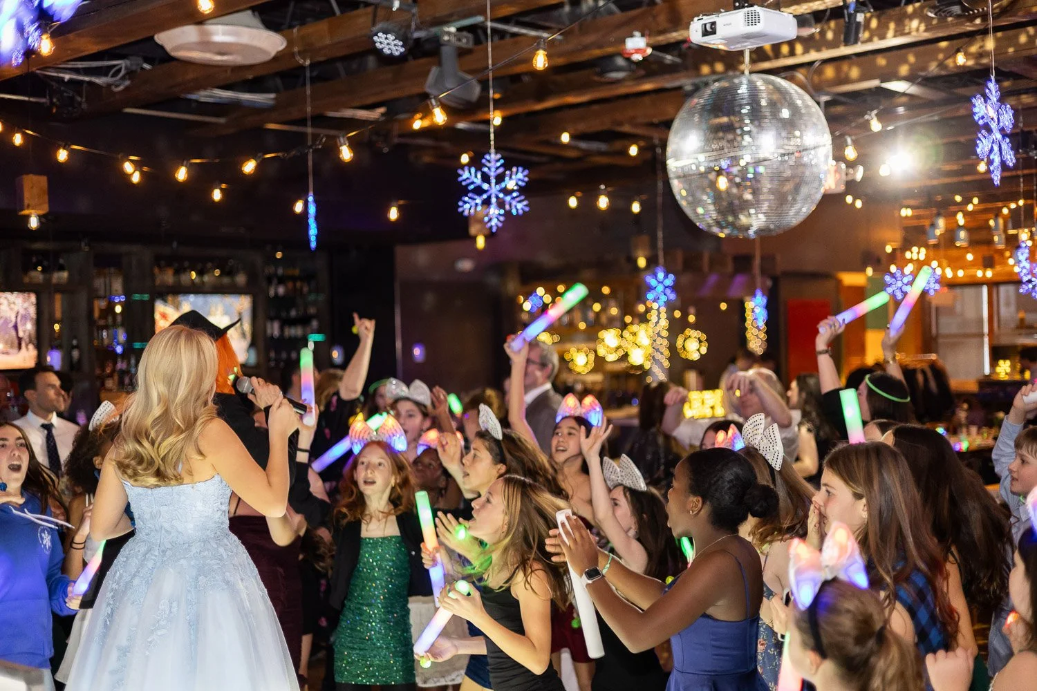 Children and adults dancing and celebrating at a Bar Mitzvah with glow sticks, fairy lights, blue snowflake decorations, and a disco ball in a decorated indoor venue.
