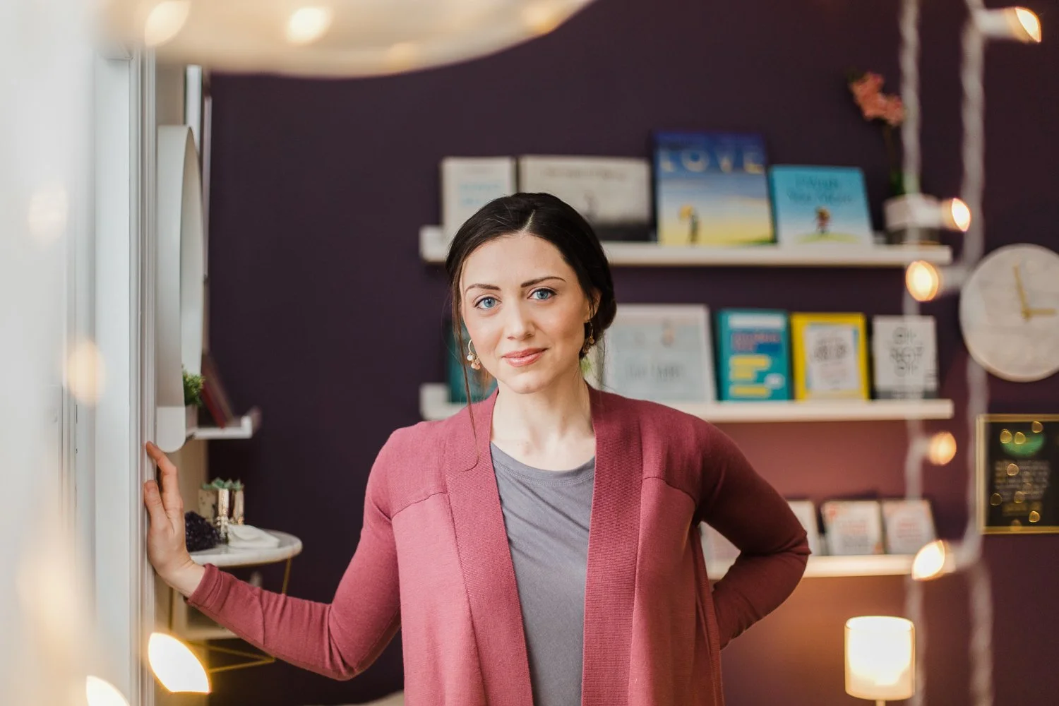 A woman in a pink blazer standing in a room with dark purple walls, bookshelves, and decorative lights.