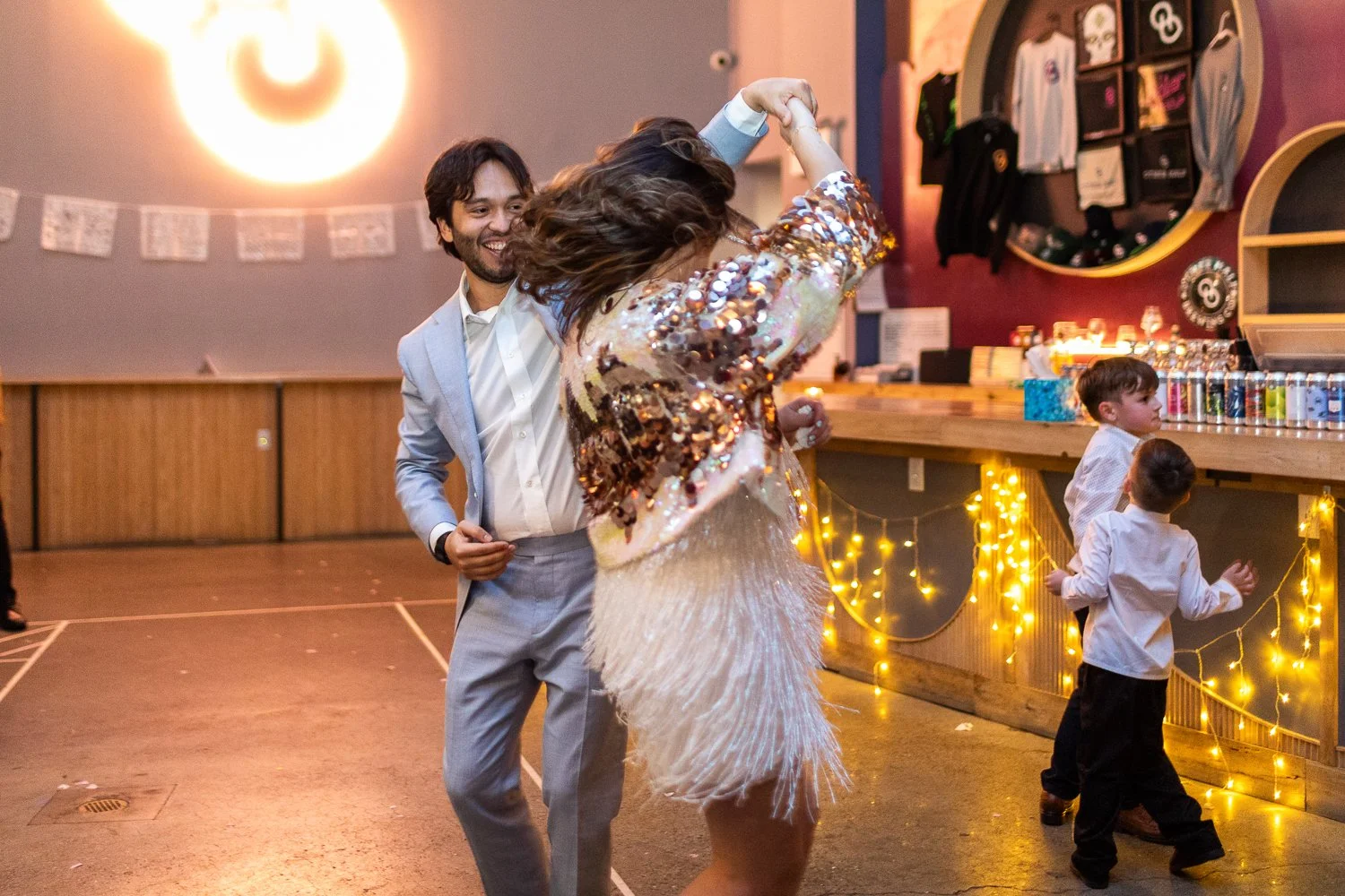 A man and woman dancing at a party, with two children nearby, and colorful decorations and fairy lights in the background.