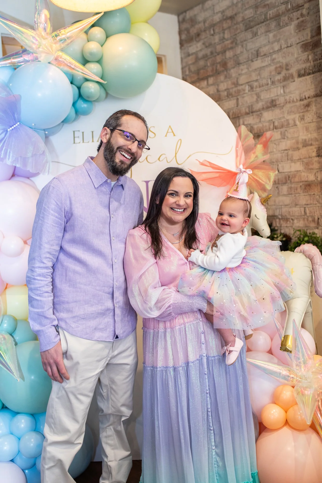 A woman holding a smiling baby girl dressed as a unicorn at a pastel-themed birthday party, with a man standing beside them, in front of a balloon and decoration backdrop with a unicorn figure.