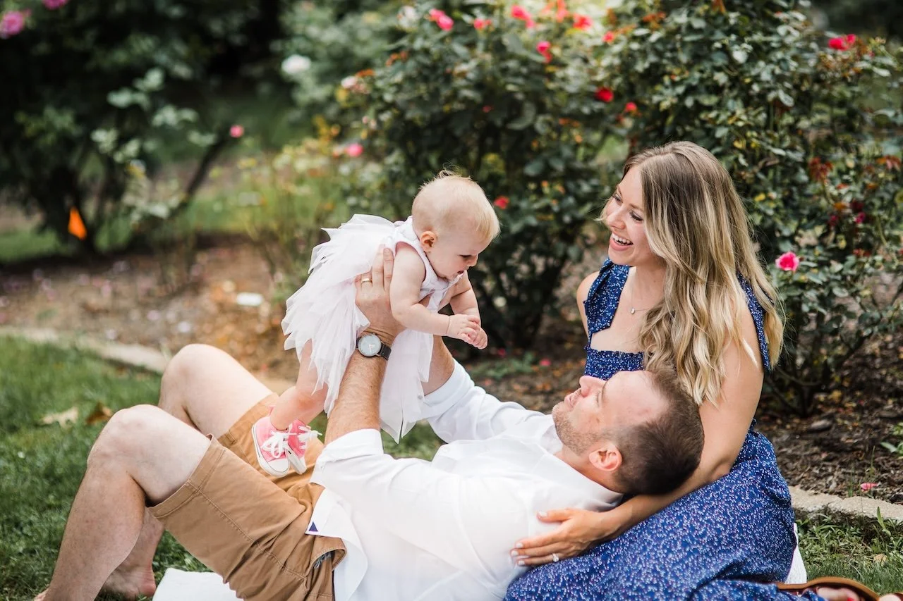 A happy family lying on the grass in a garden, playing together, with pink flowers and greenery in the background.