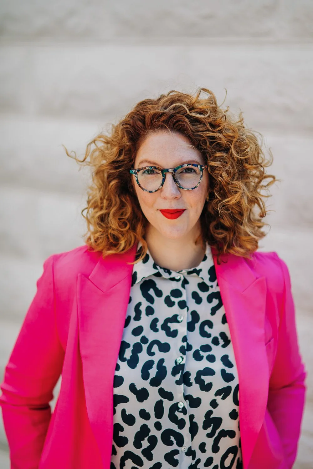 Woman with curly red hair wearing glasses, red lipstick, a leopard print shirt, and a bright pink blazer, standing in front of a light-colored wall.