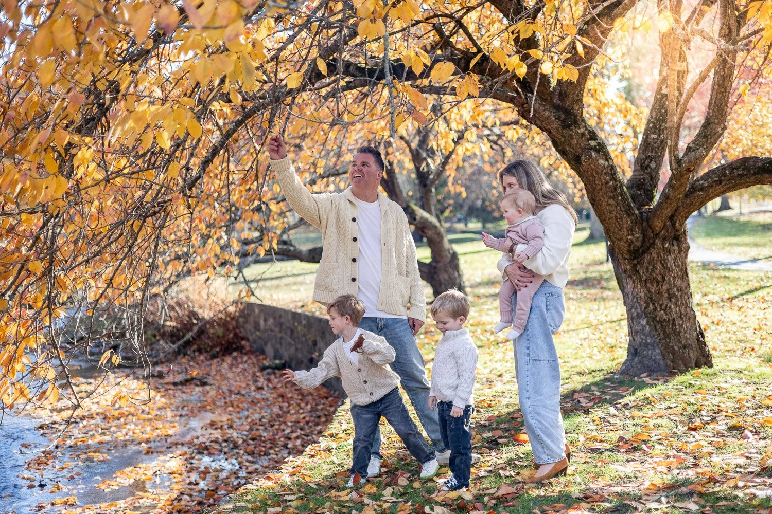 Family enjoying fall outdoors. A man and a woman with three young children near a large tree with orange and yellow leaves; the man is pointing at the leaves, and the children are playing on the ground.