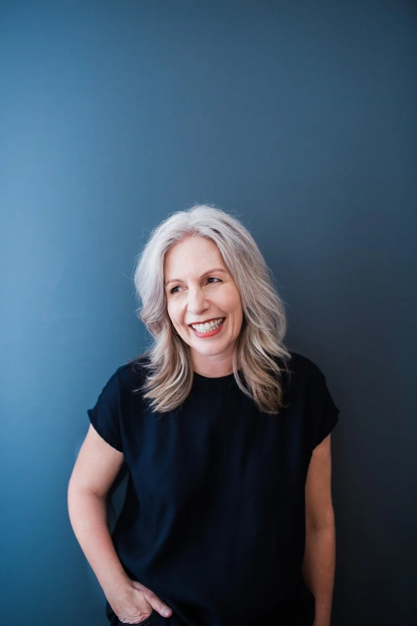 A middle-aged woman with long gray hair, smiling and looking to her right, standing against a solid blue background, wearing a black shirt.