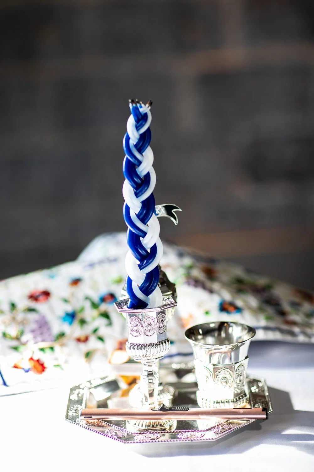 Close-up of a twisted blue and white candle in a silver candle holder on a decorative tray, with other silver items and a colorful fabric background.