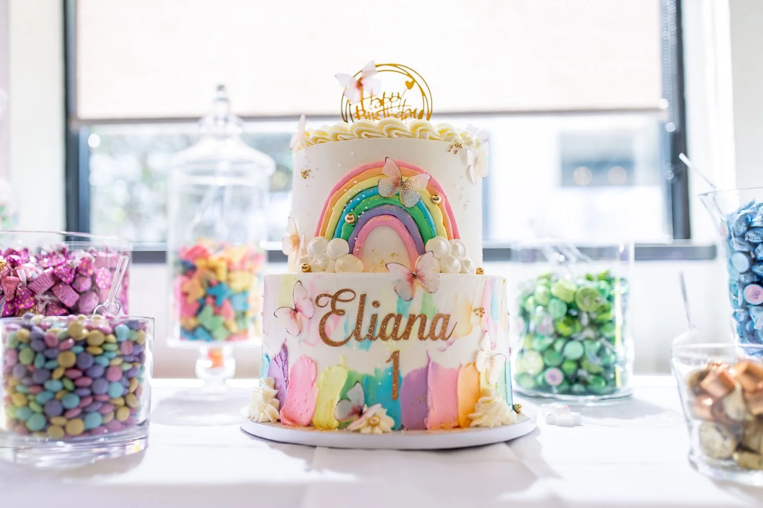 A colorful 1st birthday cake decorated with a rainbow, butterflies, and the name 'Eliana' with the number '1', surrounded by jars of candies on a table near a window.  1st Birthday Party in NJ.