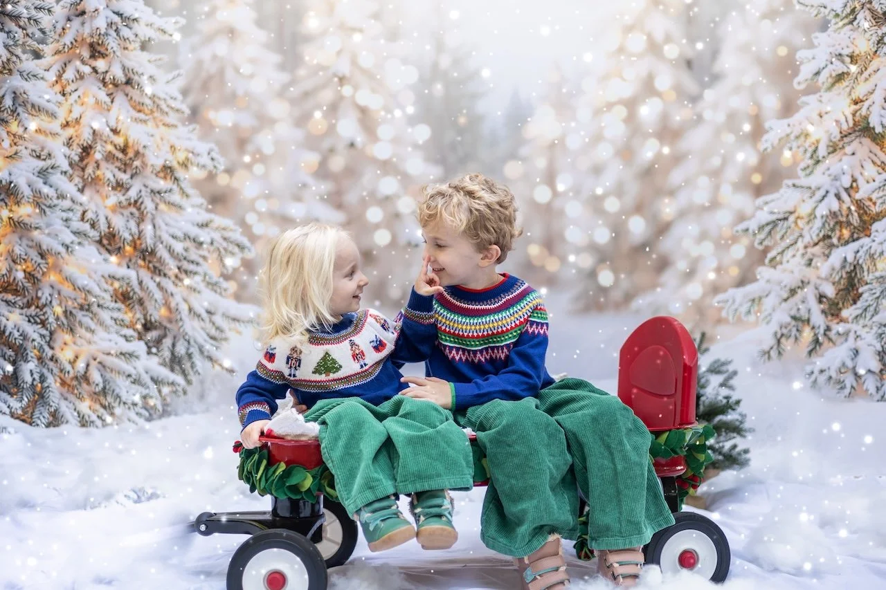 Two children, a boy and a girl, sitting on a red wagon in a snowy winter forest, wearing festive sweaters, smiling, with snow-covered trees in the background.