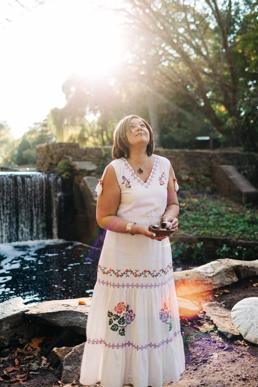 Woman in a white embroidered dress standing by a waterfall outdoors, holding small bowls, with sunlight and trees in the background.