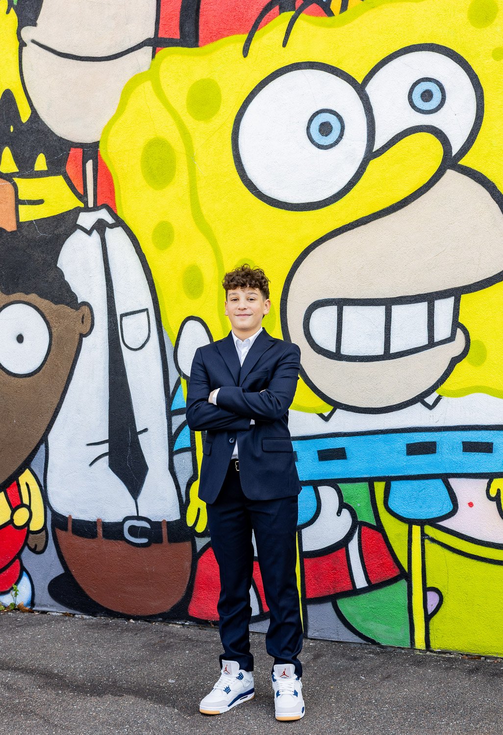 A young man in a navy suit and white sneakers standing with arms crossed in front of a colorful mural featuring characters from SpongeBob SquarePants and other cartoons.