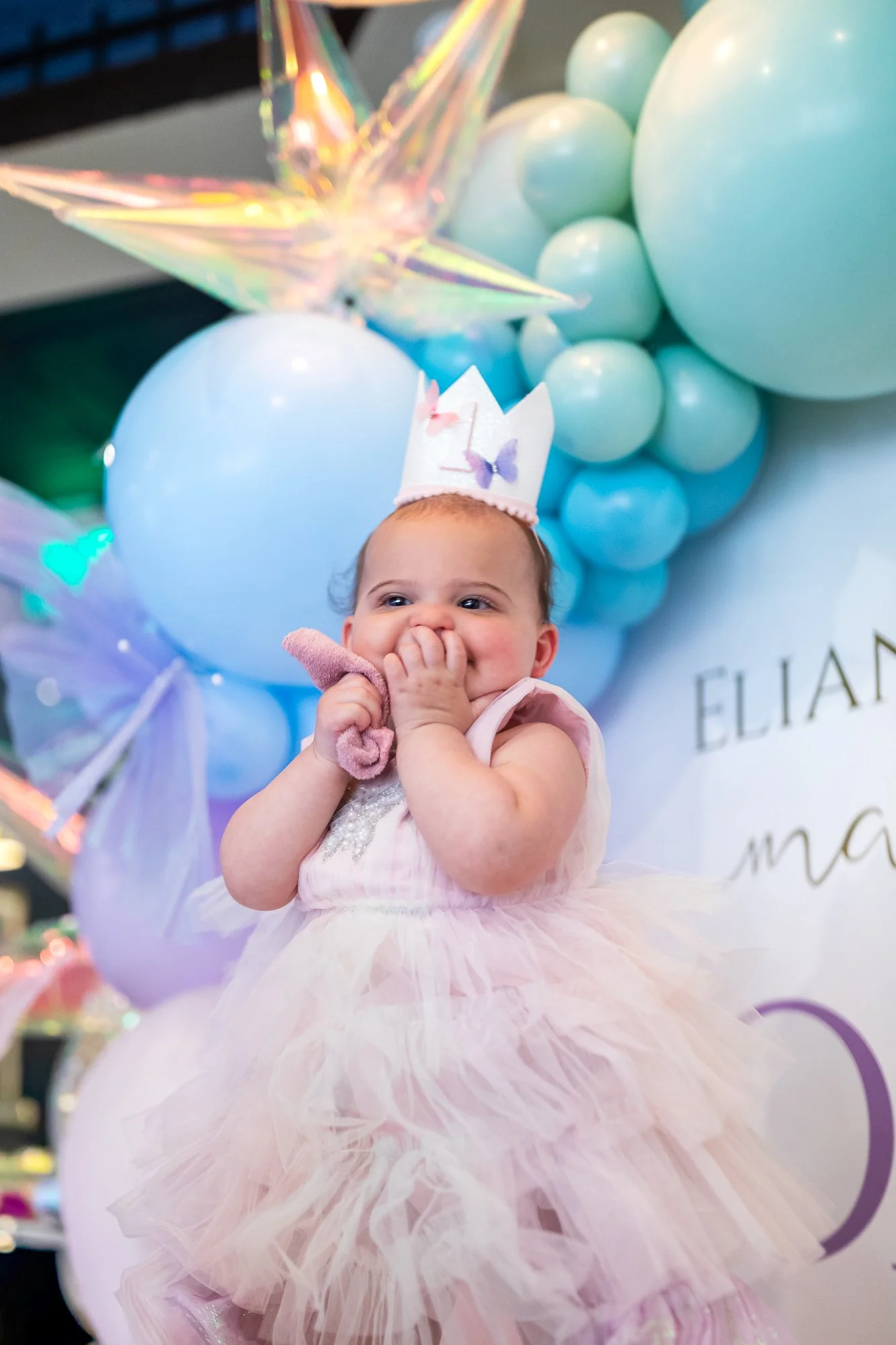 A young girl wearing a pink and white tulle dress, a butterfly headband, and holding a pink cloth, standing in front of blue and green balloons and iridescent decorations, with a background of balloons and decorations, at her 1st birthday party
.