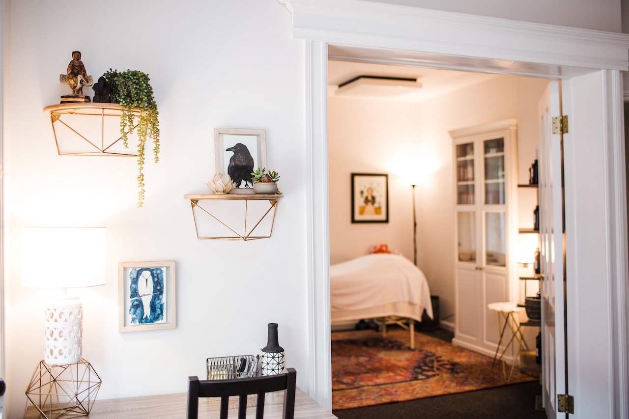 Interior view of a cozy living space with a partially visible bedroom, decorative items, wall shelves with plants and figurines, a table lamp, framed artwork, and a colorful rug.