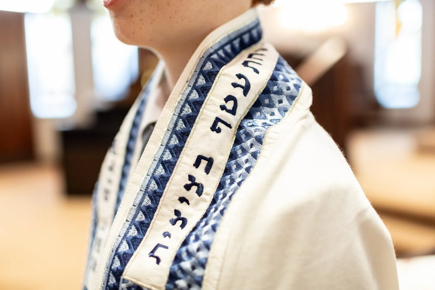 Close-up of a young boy wearing a tallit, a Jewish prayer shawl with blue and white patterns and Hebrew writing along the edge, inside a NJ synagogue during his Bar Mitzvah.